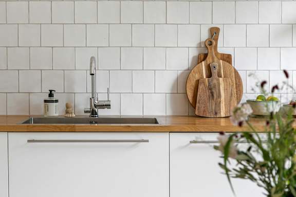 A kitchen with a white tile backsplash and a wooden cutting board on the counter.