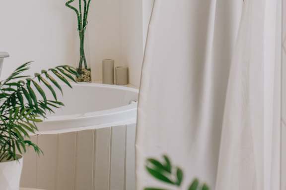 A white vase with a green plant sits on a white shelf next to a white curtain.
