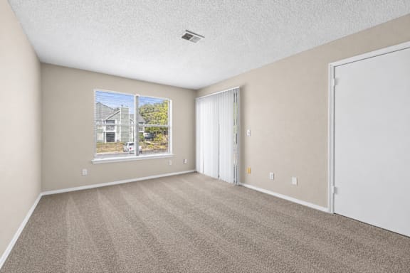 A room with a carpeted floor, a window with a view of a building and trees outside, and a white door.