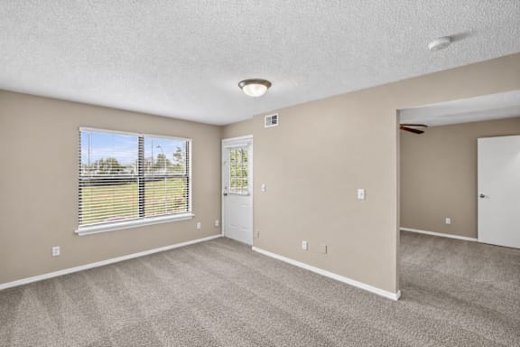A room with a carpeted floor, a window with blinds, and a door leading to another room.