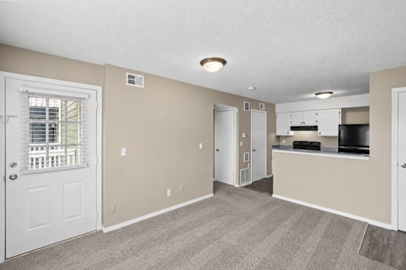 A spacious room with a window, carpeted floor, and a kitchenette in the background.