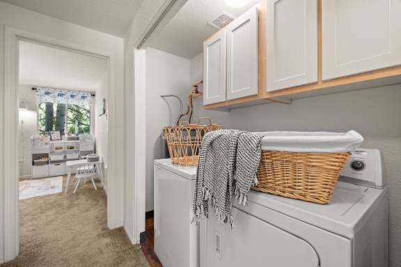 A laundry room with a washer and dryer, a basket on top of the dryer, and a window with a view of trees at Clackamas Trails Apartments, Oregon, 97222