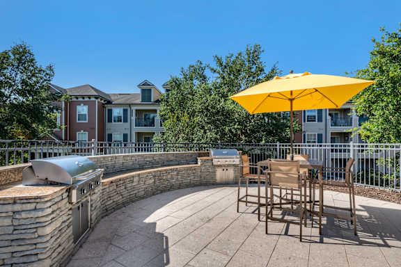 A yellow umbrella is on a patio with a stone wall and a grill.
