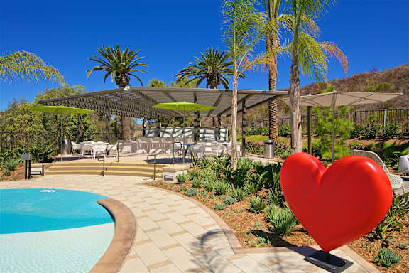 A red heart sculpture is in the foreground of a pool area with palm trees.
