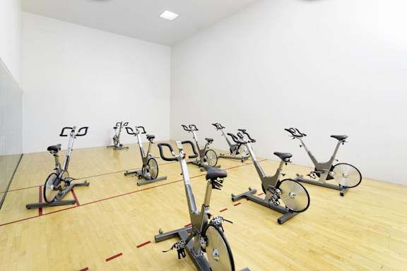 A room with a wooden floor and a white wall has several stationary bikes.