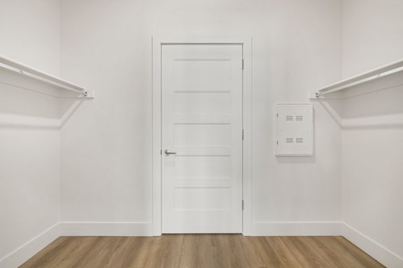 A white door is in the middle of a room with white walls and wood flooring.