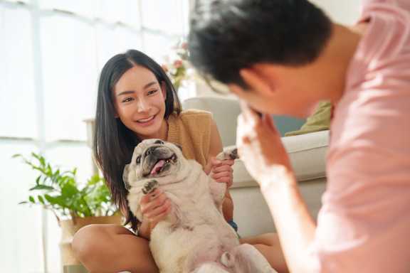 A woman is sitting on a couch with a dog, and a man is standing beside her holding a remote control.