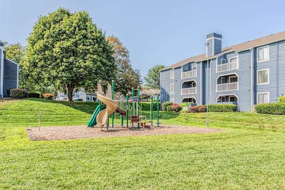 A playground with a slide and a green tree in front of a building.