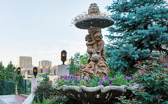 A garden with a fountain and flowers in the foreground with a city skyline in the background.