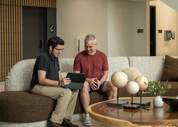 Two men are sitting on a couch, one is using a laptop.