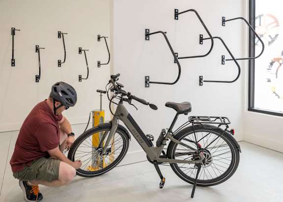 A man in a red shirt and grey shorts is fixing a bike in a room with a white wall and a black bike rack.