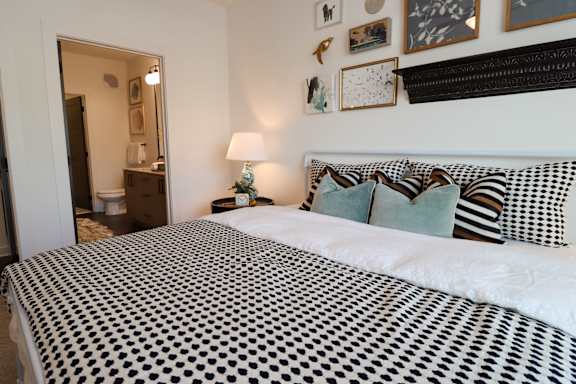 A bed with a black and white patterned cover sits in a room with a lamp and pictures on the wall.