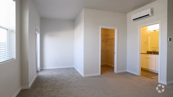 A spacious, empty room with a window on the left and a door on the right leading to a kitchen.