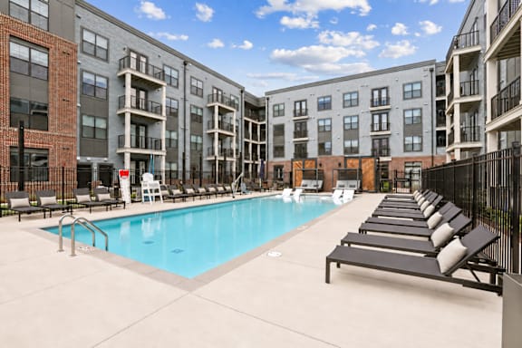 A swimming pool is surrounded by lounge chairs in front of apartment buildings.