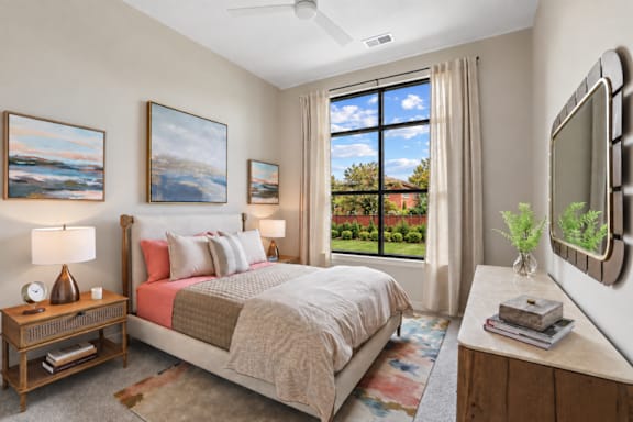 A bedroom with a large bed, a ceiling fan, and a view of the backyard through the window.