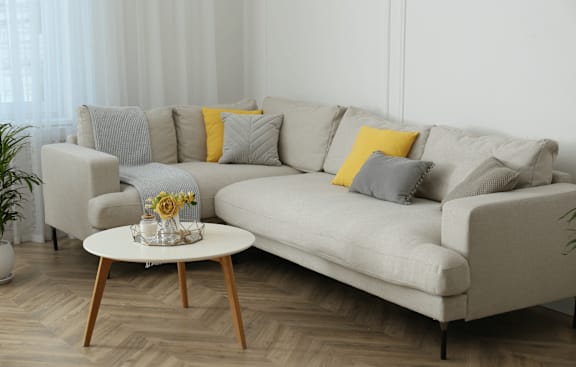 A grey couch with a yellow pillow and a small table with a vase of flowers on it.