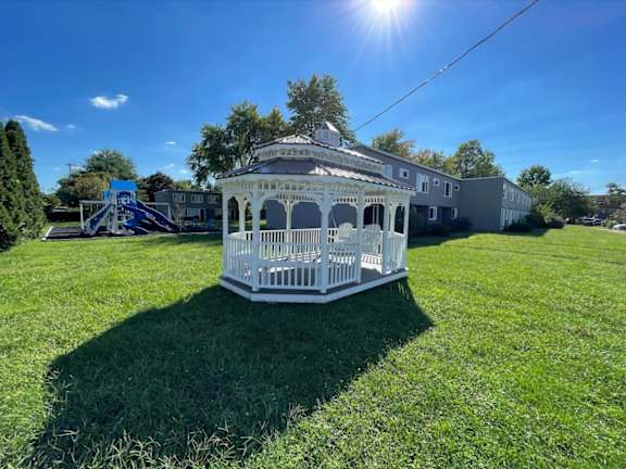 A white gazebo is situated in a grassy field with a building in the background.