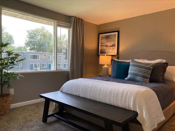 A bedroom with a bed, a bench, and a window overlooking a building.