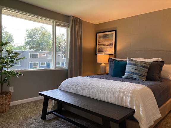 A bedroom with a bed, a bench, and a window overlooking a building.