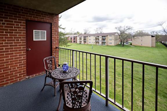 A balcony with a table and chairs overlooking a grassy area at Rockdale Gardens Apartments in Windsor Mill, MD.