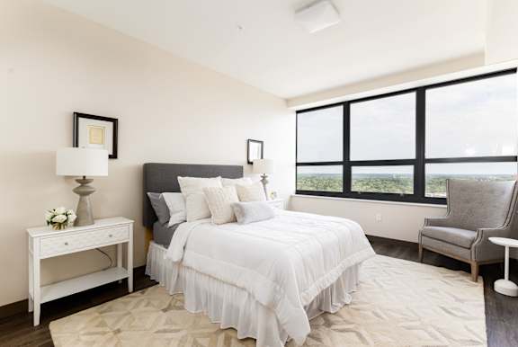 A bedroom with a large bed, a nightstand with a lamp, a chair, and a window with a view.