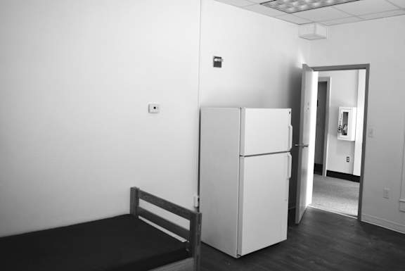 A black and white photo of a bed, a fridge and a doorway.