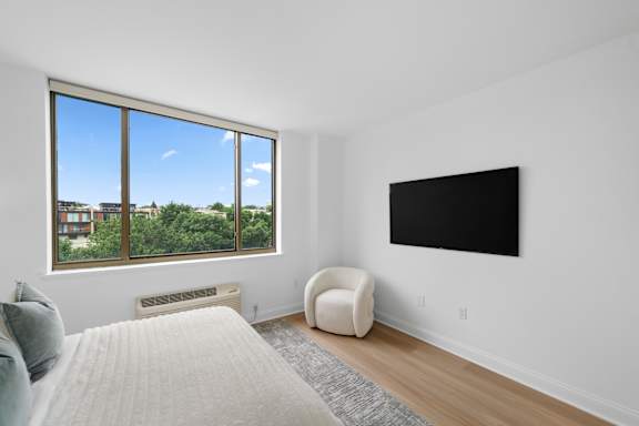 A bedroom with a large window, a bed, a chair, and a flat screen TV.