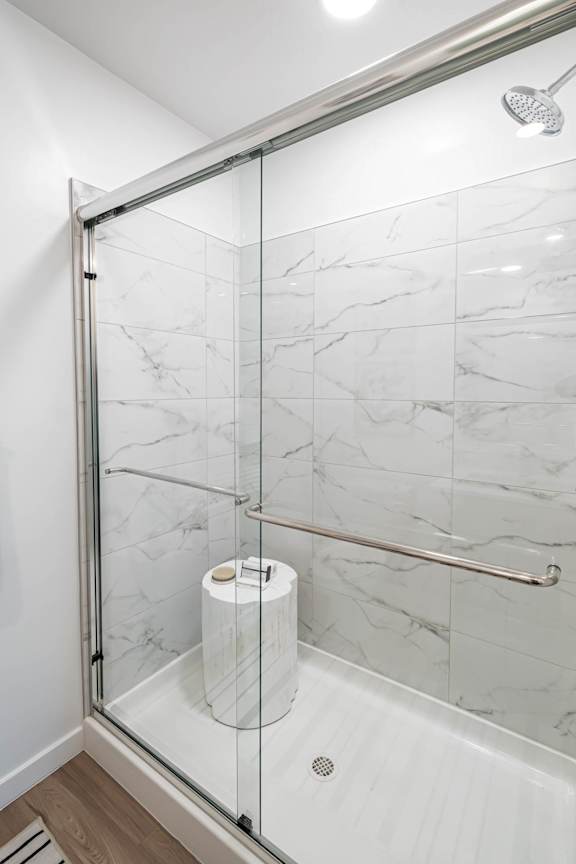 A white marble tiled shower with a glass door and a toilet paper roll on the floor.