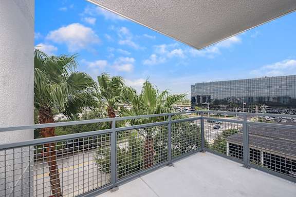 A balcony with a metal railing overlooks a parking lot and palm trees.