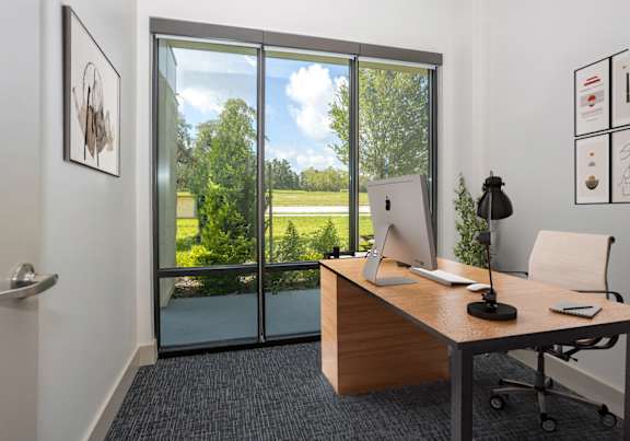 A modern office with a wooden desk and a view of the outdoors through the glass doors.