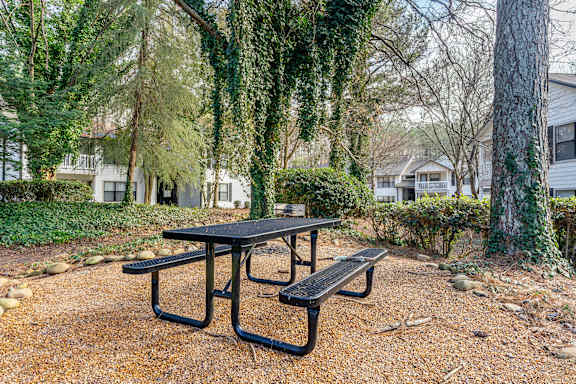A black picnic table sits in a gravel area with a tree and a house in the background.