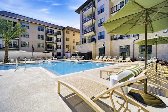 A pool area with sun loungers and umbrellas in front of apartment buildings.