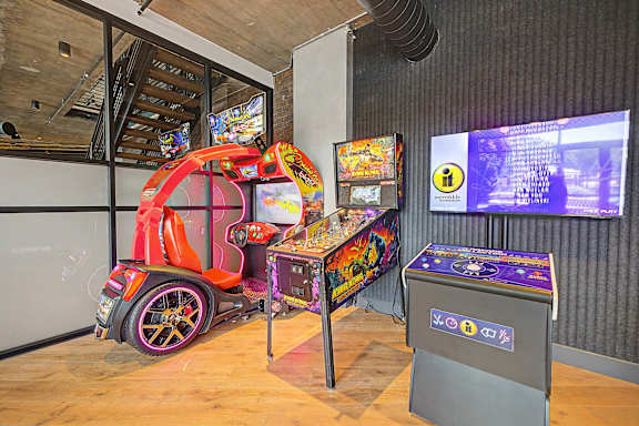 A red and black car-themed arcade game machine sits in a room with a wooden floor and a grey trash bin.