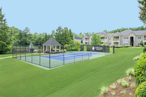 A tennis court is surrounded by a green lawn and a fence.