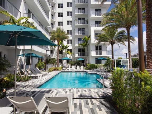 a large swimming pool with chaise lounge chairs and umbrellas in front of a white at Saba Pompano Beach, Pompano Beach Florida
