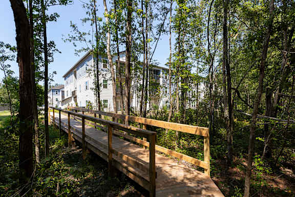 A wooden bridge leads to a white house in a wooded area.
