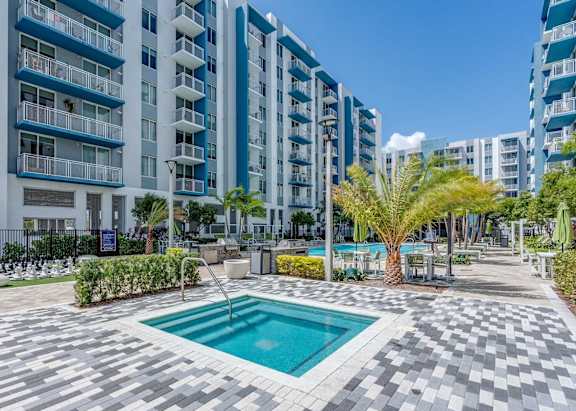 A swimming pool in the middle of a tiled area surrounded by palm trees and bushes.