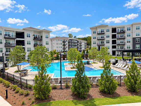A large swimming pool surrounded by a black fence and trees in front of apartment buildings.