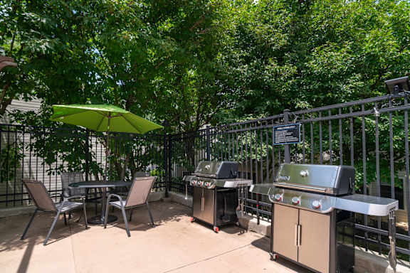 A patio with a grill and chairs under a green umbrella.