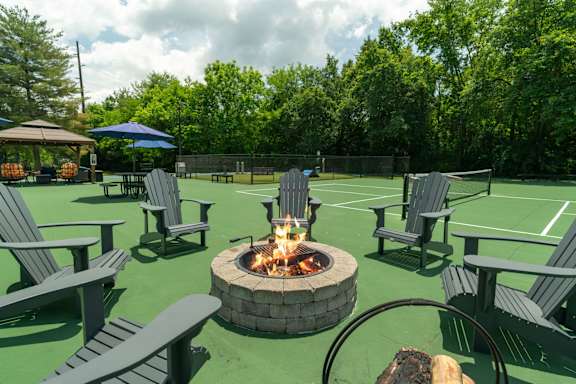 A fire pit is surrounded by Adirondack chairs on a tennis court.