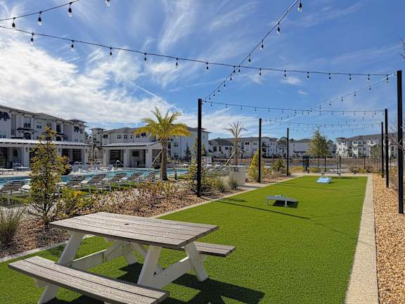 A picnic table is in the foreground of a grassy area with a pool and buildings in the background.