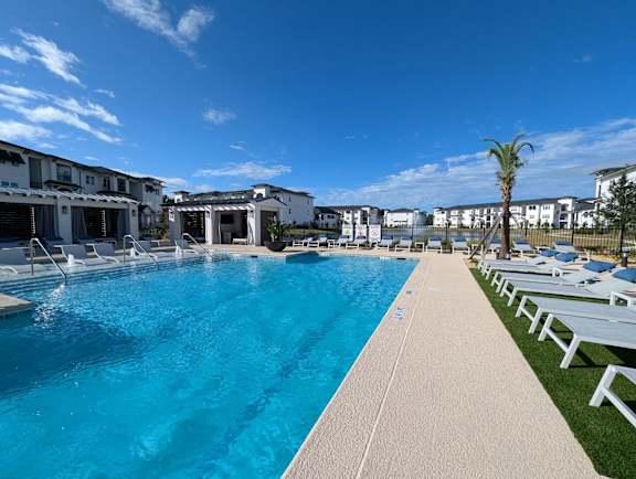 A large swimming pool with a clear blue water surrounded by white lounge chairs and buildings in the background.