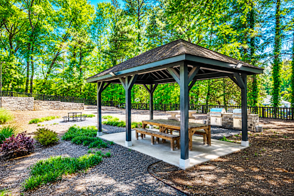 A gazebo with a thatched roof is surrounded by trees and has picnic tables.
