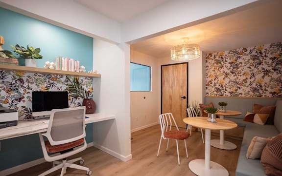 A room with a desk, chairs, and a couch with a floral wallpaper.
