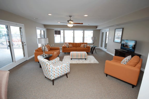 Clubhouse room with orange chairs, brown carpet and beige walls.  TV to the right of photo on wall.