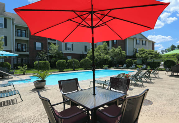 Table with 4 chairs and red umbrella by blue pool surrounded with lounge chairs.
