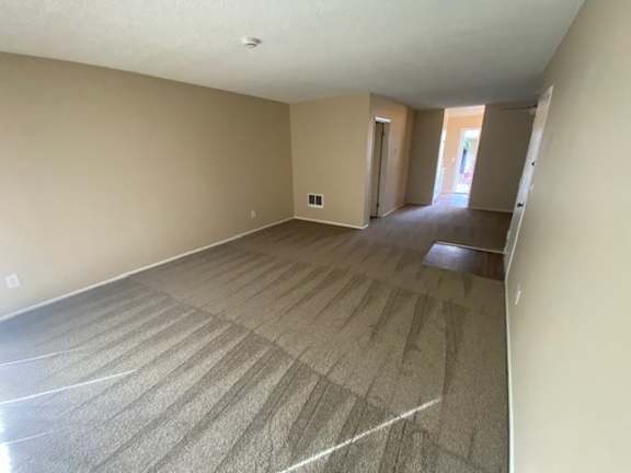 A living room with carpeted flooring and front doorway leading to another room.