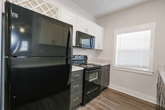 A black refrigerator is in a kitchen with white cabinets and a window with blinds at McHenry Square Apartments, Augusta, GA