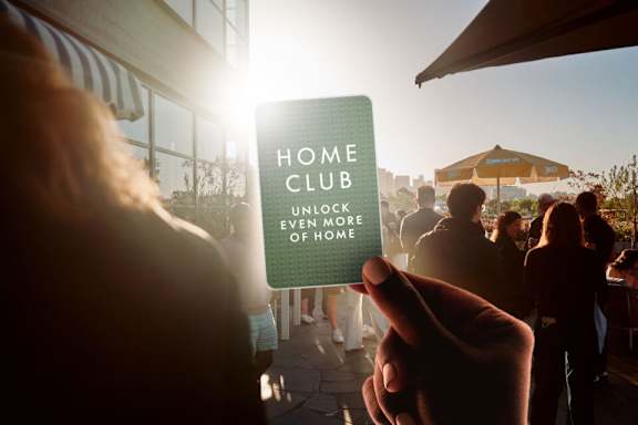 A person holding a card that says "Home Club" in front of a sunny outdoor scene.