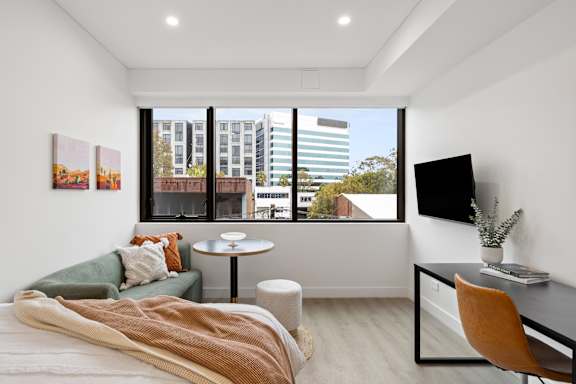 A bedroom with a bed, desk, and chair with a window overlooking a cityscape.